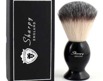 May include: A black shaving brush with a rounded handle and white bristles featuring dark tips. The brush is positioned next to a rectangular black box with a silver border, the text "Sharpy ENGLAND," and a shield logo containing a lion head graphic.