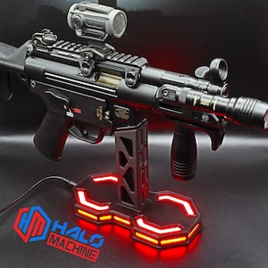 May include: A black firearm with a mounted scope and flashlight, resting on a custom stand with red LED lighting. The stand features the text "HALO MACHINE" in red and blue. The firearm has a black grip and various attachments.