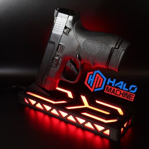 May include: Black handgun resting on a black base with red LED lighting. The base has a geometric design and the text "HALO MACHINE" in blue and red. The handgun is angled upwards.