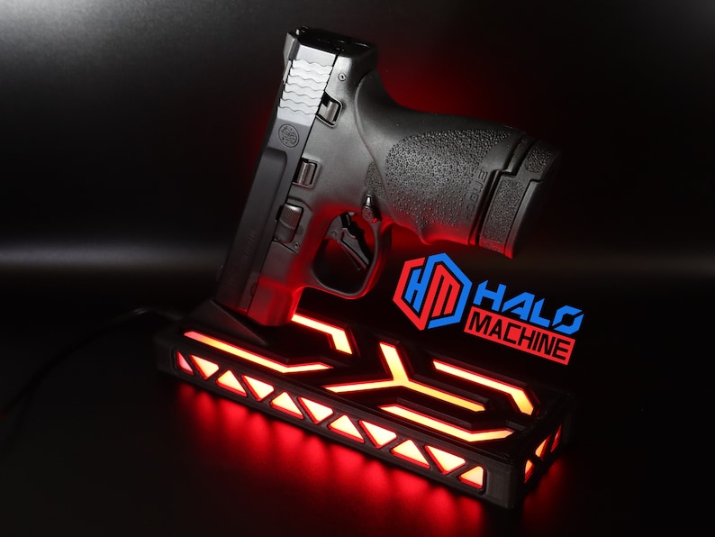 May include: Black handgun resting on a black base with red LED lighting. The base has a geometric design and the text "HALO MACHINE" in blue and red. The handgun is angled upwards.