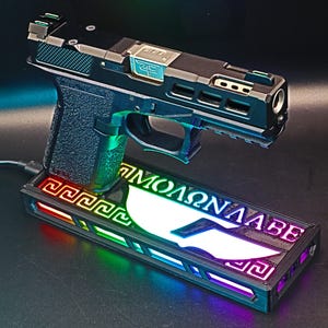May include: A black and silver handgun with a green grip rests on a rectangular base. The base has a Greek key design and the words "&Mu;&Omicron;&Lambda;&Omega;&Nu; &Lambda;&Alpha;&Beta;&Epsilon;" in white. The base is illuminated with a rainbow of colors.