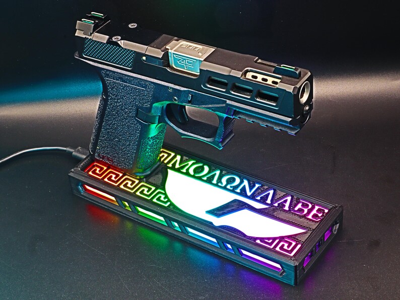 May include: A black and silver handgun with a green grip rests on a rectangular base. The base has a Greek key design and the words "&Mu;&Omicron;&Lambda;&Omega;&Nu; &Lambda;&Alpha;&Beta;&Epsilon;" in white. The base is illuminated with a rainbow of colors.