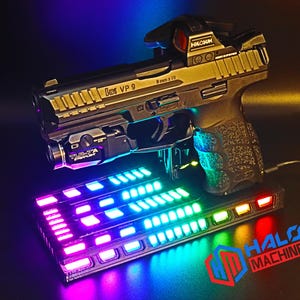 May include: A black and gold handgun with a red dot sight and a laser sight, resting on a base with colorful LED lights. The base has the text "HALO MACHINE" in red and blue.