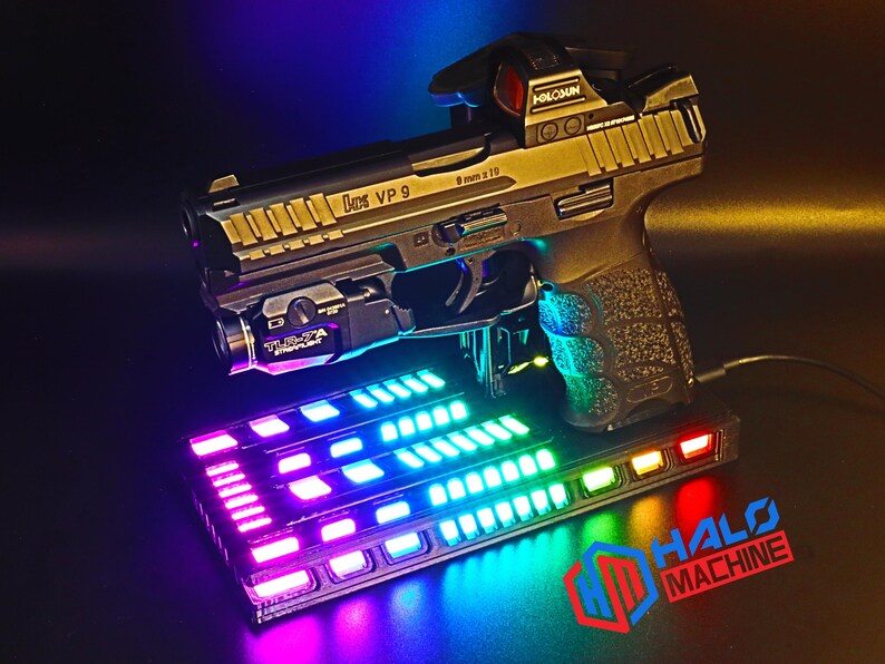 May include: A black and gold handgun with a red dot sight and a laser sight, resting on a base with colorful LED lights. The base has the text "HALO MACHINE" in red and blue.