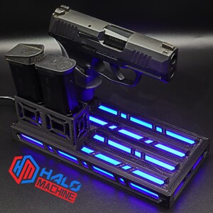 May include: A black handgun rests on a black base with blue LED lights. Two black magazines are in a holder. The base has the text "HALO MACHINE" and a logo in red and blue.