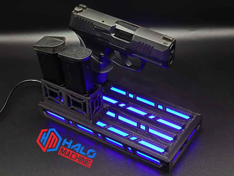 May include: A black handgun rests on a black base with blue LED lights. Two black magazines are in a holder. The base has the text "HALO MACHINE" and a logo in red and blue.