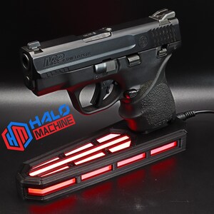 May include: A black handgun, labeled "M&P Shield Plus," rests on a black base with red illuminated stripes. The base has the text "HALO MACHINE" and a logo. A black cord extends from the base.