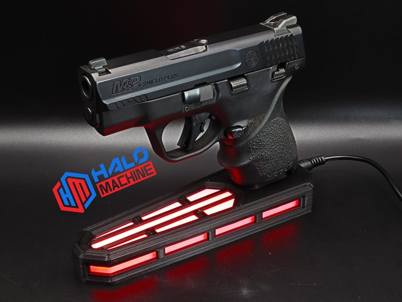 May include: A black handgun, labeled "M&P Shield Plus," rests on a black base with red illuminated stripes. The base has the text "HALO MACHINE" and a logo. A black cord extends from the base.
