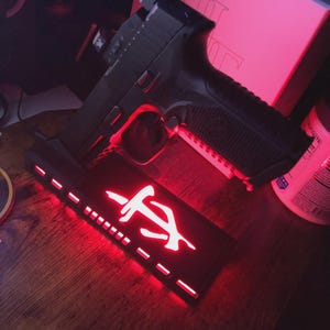 May include: A black handgun rests on a black stand with a red illuminated logo. The stand has a series of small rectangular cutouts along the bottom edge. The background is dimly lit with red light.