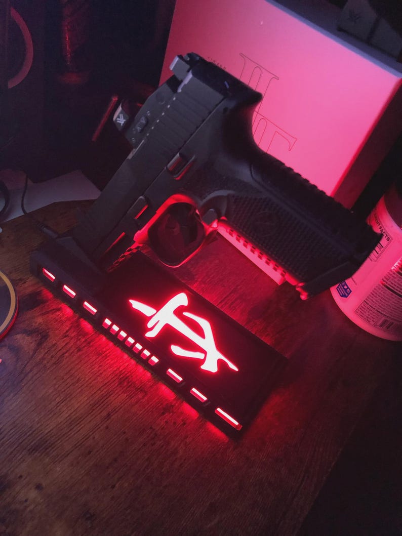 May include: A black handgun rests on a black stand with a red illuminated logo. The stand has a series of small rectangular cutouts along the bottom edge. The background is dimly lit with red light.