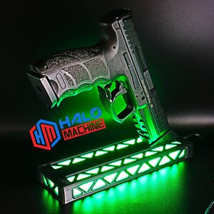 May include: A black handgun resting on a base with green LED lights. The base has a geometric design and the text "HALO MACHINE" in red, blue, and white. The gun is angled, showcasing its details against a dark background.