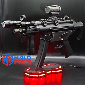 May include: Black firearm with attached accessories, including a flashlight and scope. The firearm is mounted on a black stand with red LED lighting. The Halo Machine logo is visible in the lower left corner.