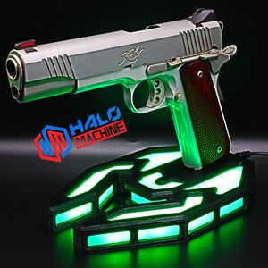 May include: A silver handgun with a wooden grip sits on a black base with green LED lights. The base has a geometric design and the text "HALO MACHINE" is visible. The handgun has a red front sight.
