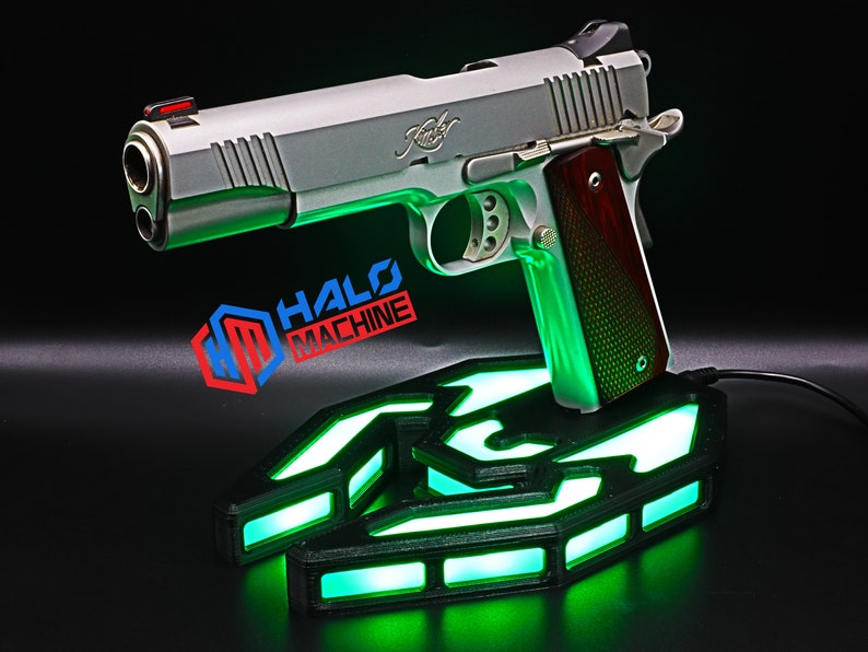 May include: A silver handgun with a wooden grip sits on a black base with green LED lights. The base has a geometric design and the text "HALO MACHINE" is visible. The handgun has a red front sight.