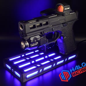 May include: Black handgun with a red dot sight and a mounted flashlight, resting on a rectangular base with blue LED lights. The base has the text "HALO MACHINE" in red and blue.