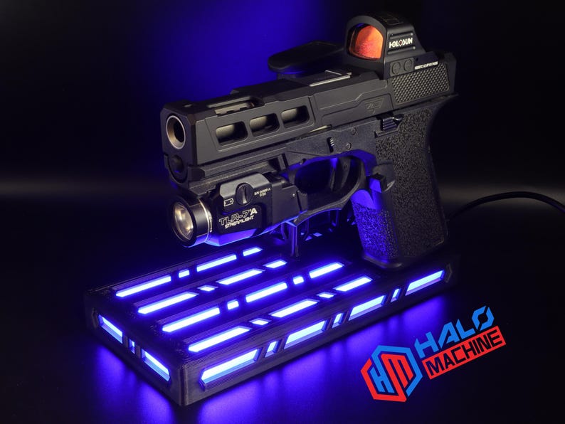 May include: Black handgun with a red dot sight and a mounted flashlight, resting on a rectangular base with blue LED lights. The base has the text "HALO MACHINE" in red and blue.