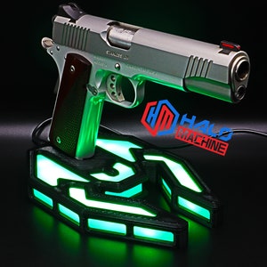 May include: A silver handgun with a brown handle is displayed on a black base with green LED illumination. The base features a geometric design, and the text "HALO MACHINE" is visible.