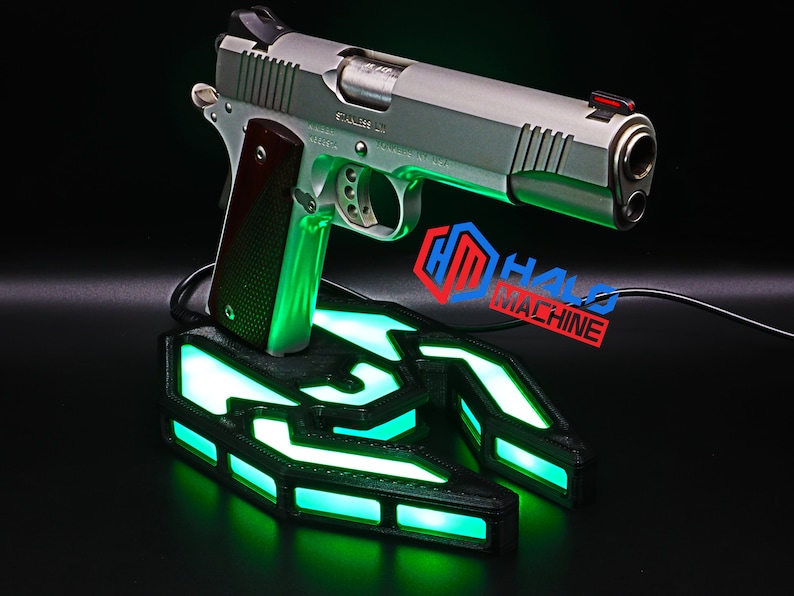 May include: A silver handgun with a brown handle is displayed on a black base with green LED illumination. The base features a geometric design, and the text "HALO MACHINE" is visible.