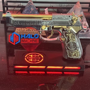 May include: A silver and gold handgun with intricate engravings, resting on a black base with red LED lighting. The text "HALO MACHINE" is visible. The gun features ornate detailing and a polished finish.