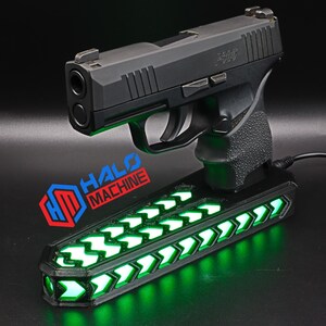 May include: A black handgun rests on a black base with green LED lights. The base has a series of green arrow-shaped lights. The text "HALO MACHINE" is visible on the base.
