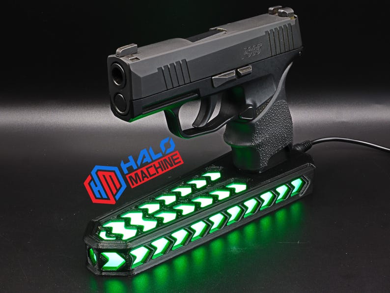 May include: A black handgun rests on a black base with green LED lights. The base has a series of green arrow-shaped lights. The text "HALO MACHINE" is visible on the base.