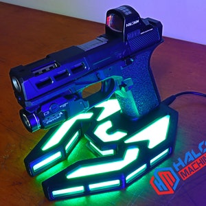 May include: A black handgun with a red dot sight and a mounted flashlight rests on a glowing green and white base. The base has a stylized design with the text "HALO MACHINE" and the initials "HM".
