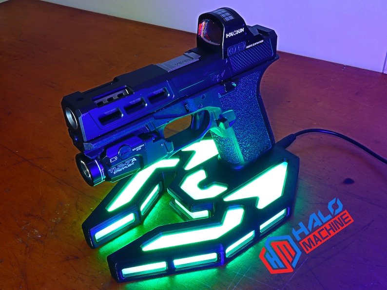 May include: A black handgun with a red dot sight and a mounted flashlight rests on a glowing green and white base. The base has a stylized design with the text "HALO MACHINE" and the initials "HM".