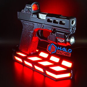 May include: A black handgun with a red dot sight and a mounted flashlight, displayed on a stand with red LED lights. The stand features the text "HALO MACHINE" in blue and red. The gun has the text "7401".