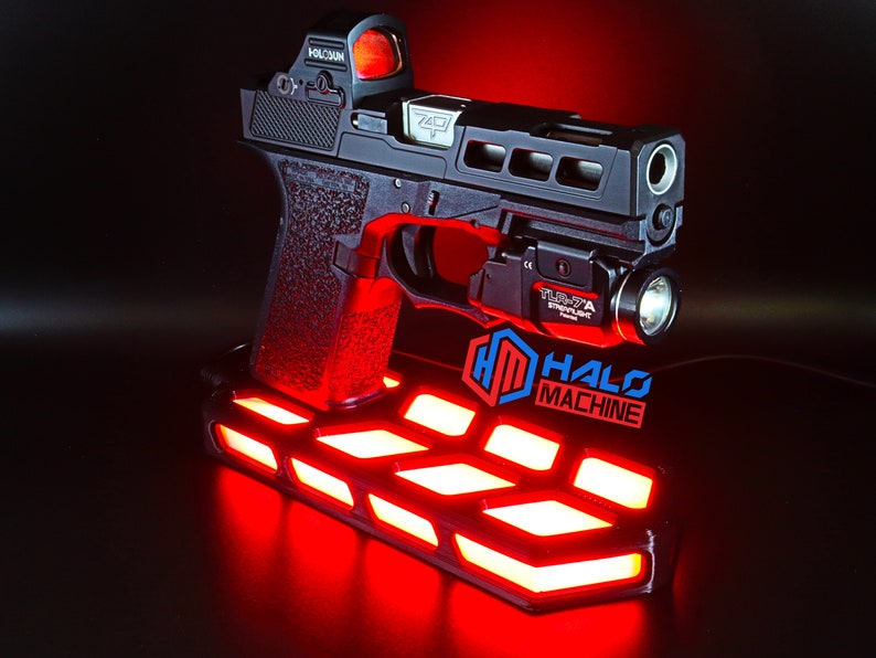May include: A black handgun with a red dot sight and a mounted flashlight, displayed on a stand with red LED lights. The stand features the text "HALO MACHINE" in blue and red. The gun has the text "7401".