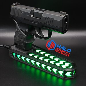 May include: A black handgun rests on a black base with green LED lights. The base has a series of chevron-shaped lights. The text "HALO MACHINE" is visible in blue and red.