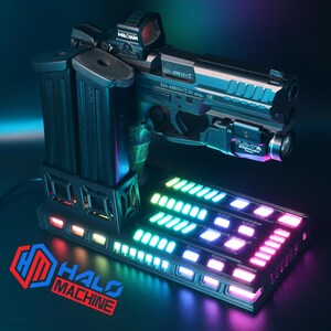 May include: A black handgun with a red dot sight and a mounted flashlight, resting on a base with a multi-colored LED display. Two black magazines are attached to the side. The base has the text "HALO MACHINE".