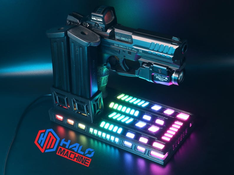 May include: A black handgun with a red dot sight and a mounted flashlight, resting on a base with a multi-colored LED display. Two black magazines are attached to the side. The base has the text "HALO MACHINE".
