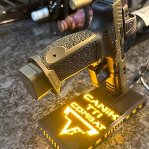 May include: A bronze and black handgun with a textured grip, resting on a black base. The base has the words "CANIK TTI COMBAT" illuminated in yellow. The handgun is angled upwards, showcasing its design.