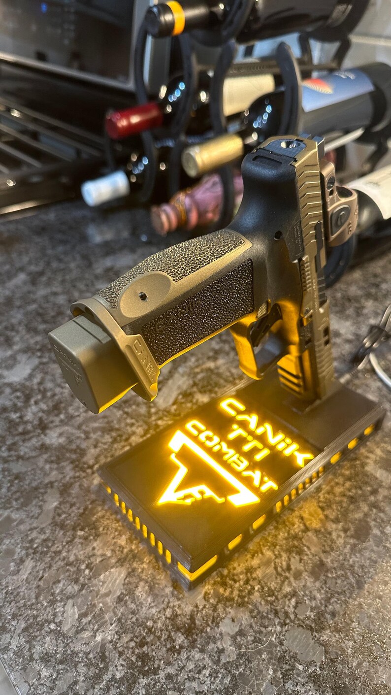 May include: A bronze and black handgun with a textured grip, resting on a black base. The base has the words "CANIK TTI COMBAT" illuminated in yellow. The handgun is angled upwards, showcasing its design.