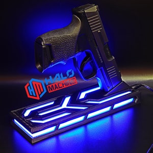 May include: Black handgun resting on a custom stand with blue LED lighting. The stand has a geometric design and the text "HALO MACHINE" in red and blue. A black cable is connected to the stand.