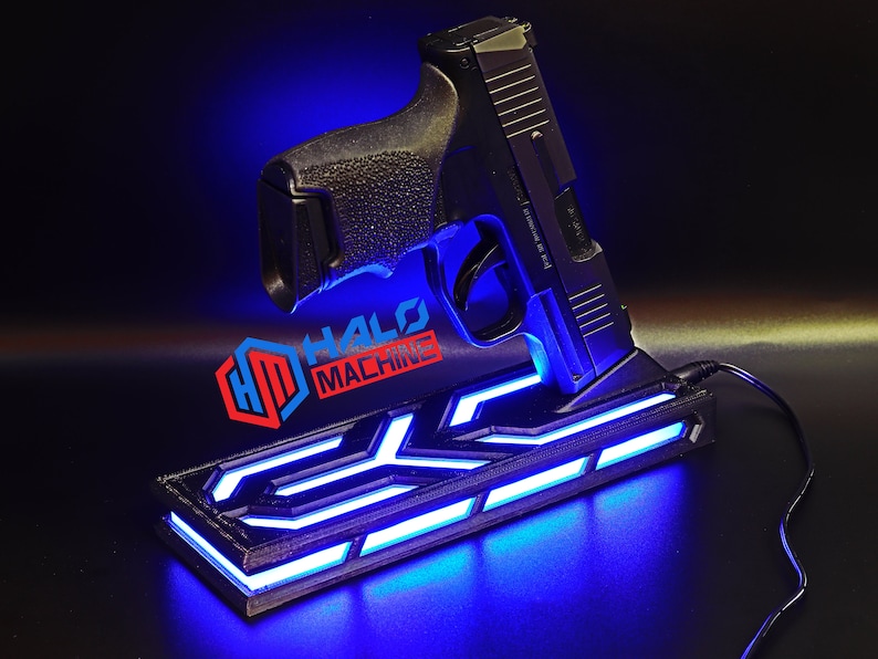 May include: Black handgun resting on a custom stand with blue LED lighting. The stand has a geometric design and the text "HALO MACHINE" in red and blue. A black cable is connected to the stand.