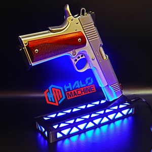 May include: A silver and wood-toned handgun displayed on a black base with blue LED lights. The base has a geometric design and the text "HALO MACHINE" is visible. The handgun has a wooden grip.