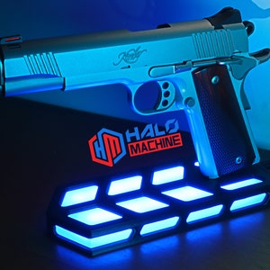 May include: A silver handgun with a wooden grip sits on a black base with blue LED lights. The base has the text "HALO MACHINE" and a logo. The handgun is a Kimber.