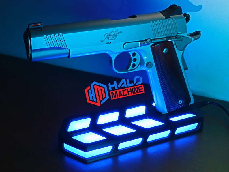 May include: A silver handgun with a wooden grip sits on a black base with blue LED lights. The base has the text "HALO MACHINE" and a logo. The handgun is a Kimber.