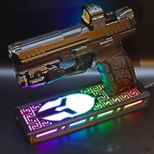 May include: A black handgun with a red dot sight and a mounted flashlight, resting on a rectangular base with a white emblem and colorful LED lighting. The gun is labeled "HK VP9".