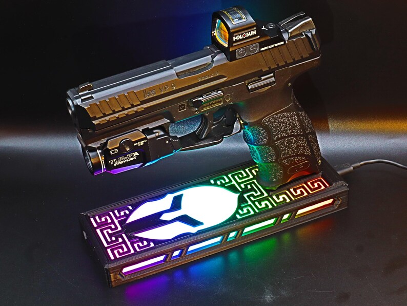 May include: A black handgun with a red dot sight and a mounted flashlight, resting on a rectangular base with a white emblem and colorful LED lighting. The gun is labeled "HK VP9".