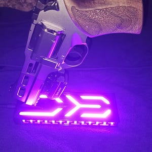 May include: A silver and black handgun with a wooden grip rests on a black base with a glowing purple arrow design. The handgun has a mounted light with the brand name "Olight".
