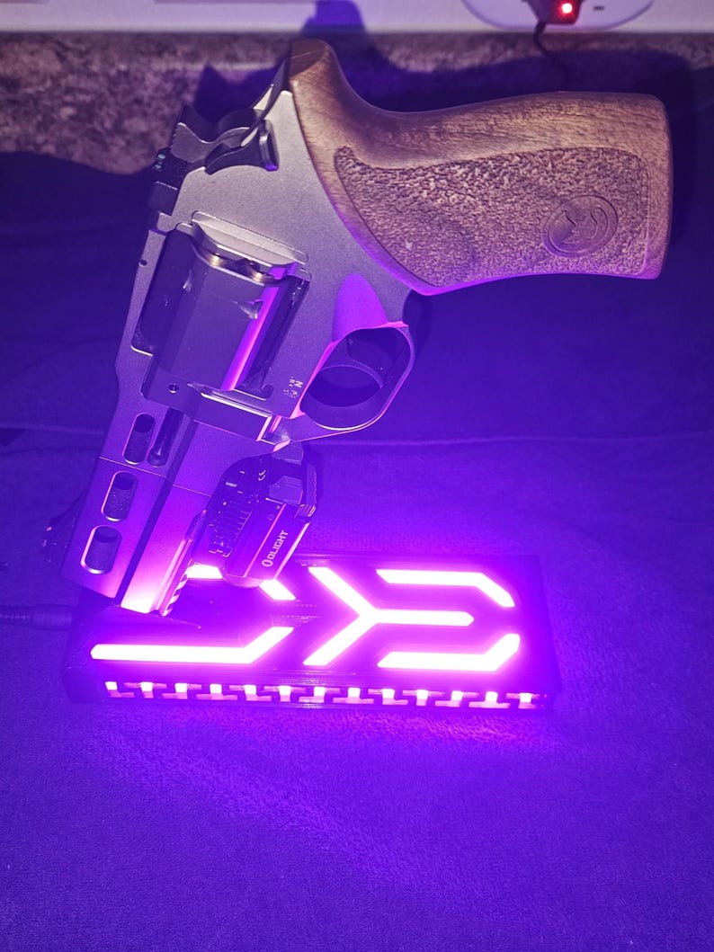 May include: A silver and black handgun with a wooden grip rests on a black base with a glowing purple arrow design. The handgun has a mounted light with the brand name "Olight".