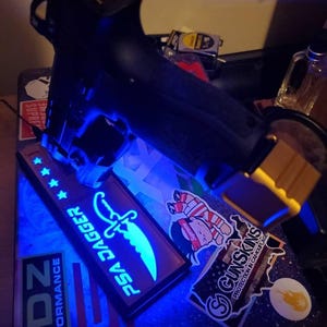May include: Black handgun with a blue light illuminating a PSA Dagger patch with a glowing knife design and text. The image also shows stickers with various designs, including the text "GUNSKINS PROTECTION IN CAMOUFLAGE."