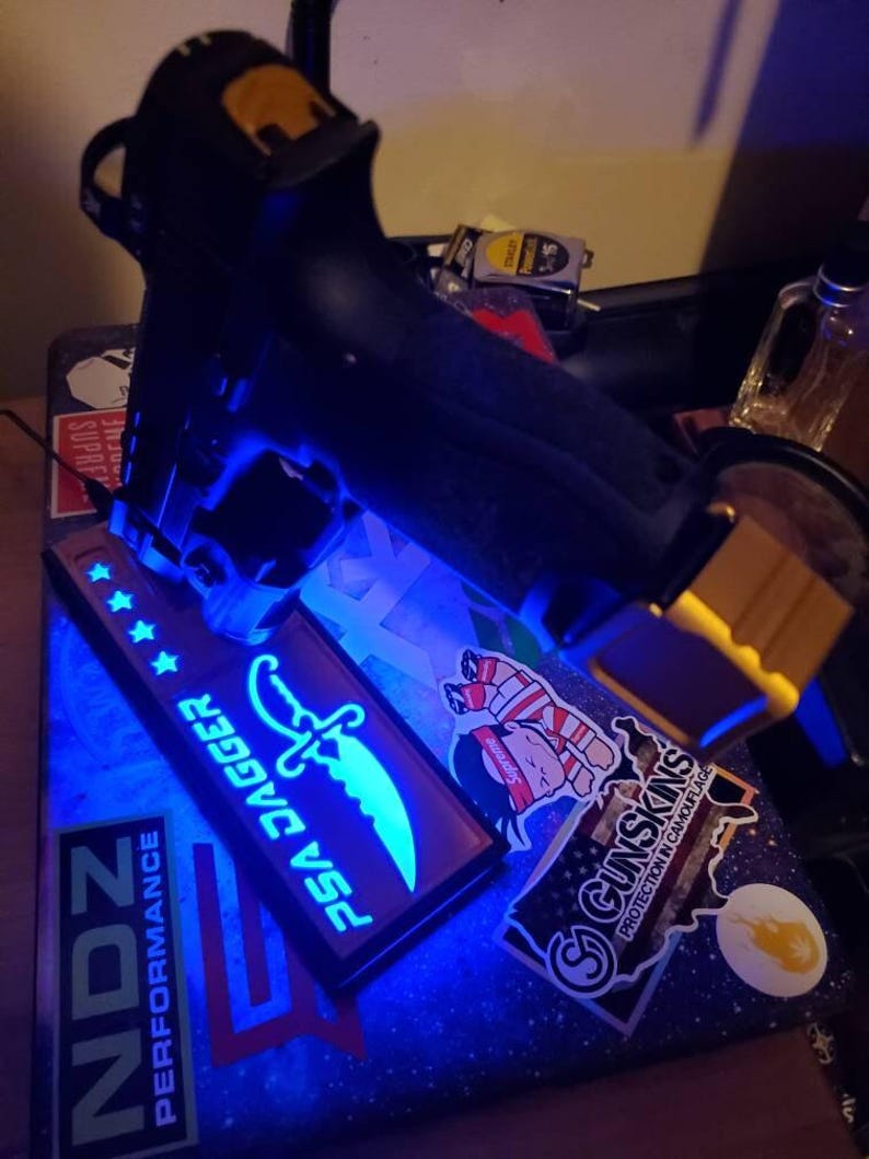 May include: Black handgun with a blue light illuminating a PSA Dagger patch with a glowing knife design and text. The image also shows stickers with various designs, including the text "GUNSKINS PROTECTION IN CAMOUFLAGE."
