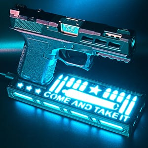 May include: A black and copper-colored handgun with a textured grip, resting above a rectangular base. The base features the text "COME AND TAKE IT" in bright blue, along with bullet and star graphics. The image is illuminated with a blue light.