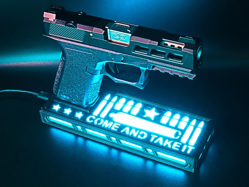 May include: A black and copper-colored handgun with a textured grip, resting above a rectangular base. The base features the text "COME AND TAKE IT" in bright blue, along with bullet and star graphics. The image is illuminated with a blue light.