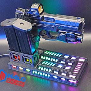 May include: A black and silver handgun with a red dot sight and a mounted flashlight rests on a black base with multi-colored LED lights. Two black magazines are in a holder on the left. The base has the text "HALO MACHINE".