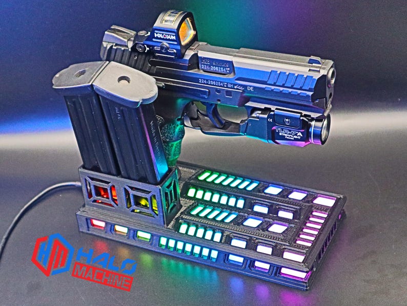 May include: A black and silver handgun with a red dot sight and a mounted flashlight rests on a black base with multi-colored LED lights. Two black magazines are in a holder on the left. The base has the text "HALO MACHINE".