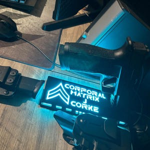 May include: A black handgun, holster, and magazine are displayed on a wooden surface. A glowing blue sign reads "CORPORAL MATRIX J CORKE" with a military rank insignia. A computer mouse and speaker are also visible.